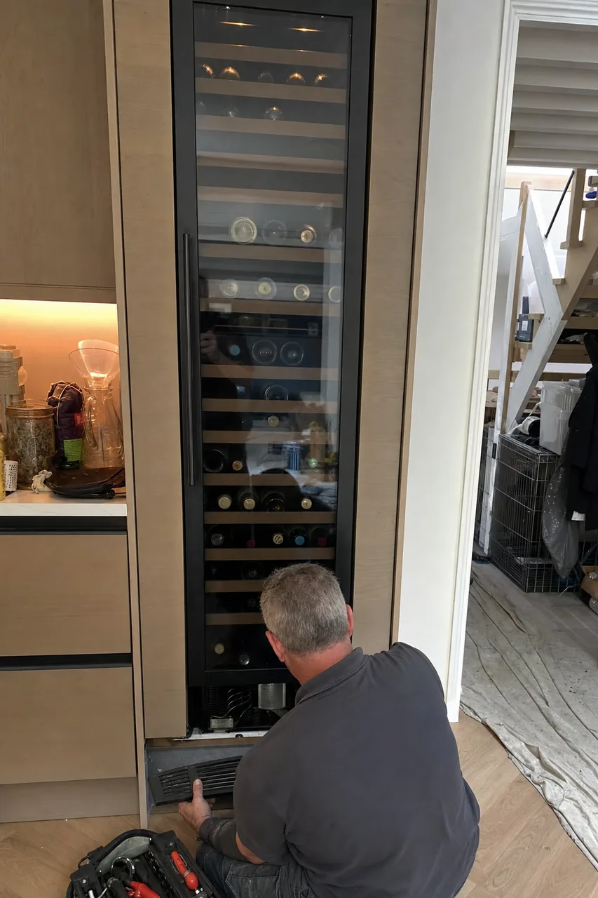 A technician in a grey work polo crouched at the base of a full-height built-in wine column, its lower grille lifted away in one hand.