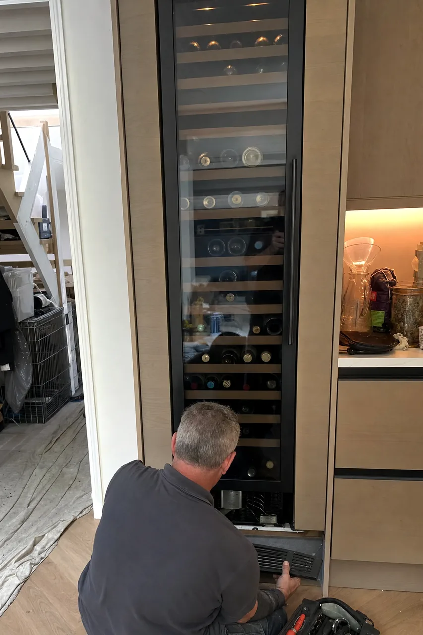 A glass-fronted refrigeration column set flush into pale wood cabinetry, bottles racked on wooden shelves behind the door.