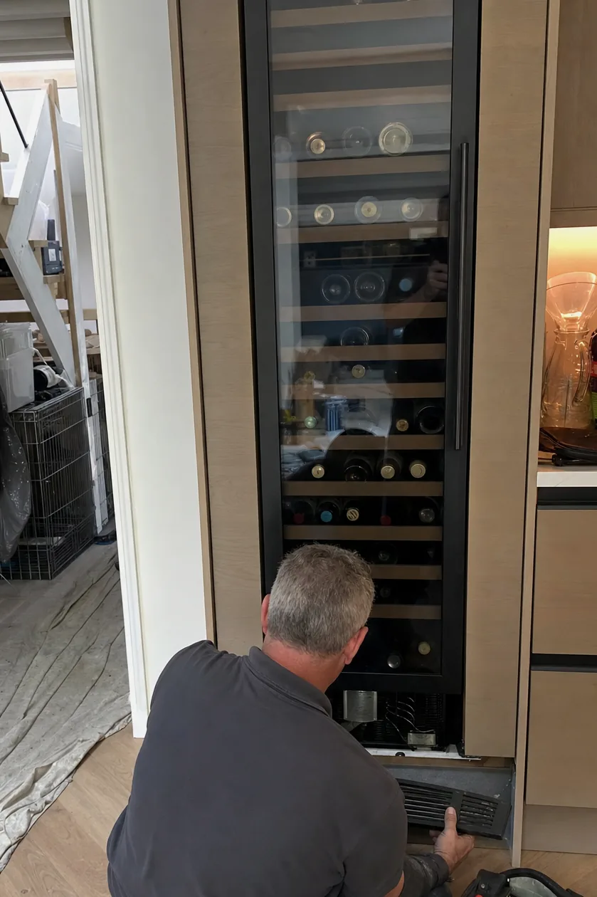 A technician kneeling at a built-in wine column in a domestic kitchen, an open tool bag on the floor beside him.