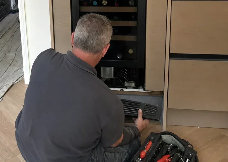 A technician kneeling at the foot of a built-in column with the lower grille held in one hand and the compressor bay open behind it, an unrolled tool kit on the floor beside him.