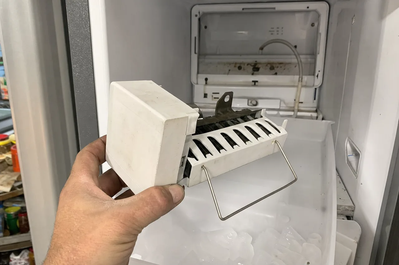 A white ice maker module with its wire harvest arm, held in one hand in front of the open freezer it came out of.