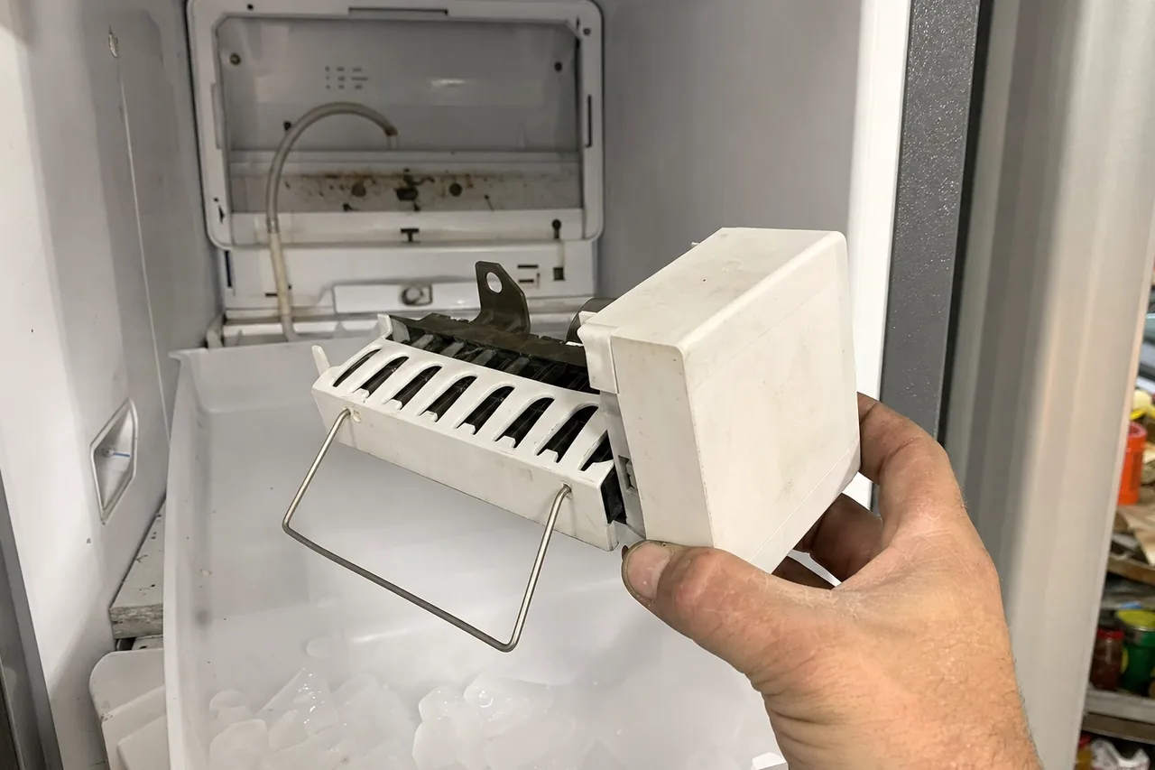 An ice maker module lifted clear of its bracket, the curved plastic fill tube visible at the back of the freezer.