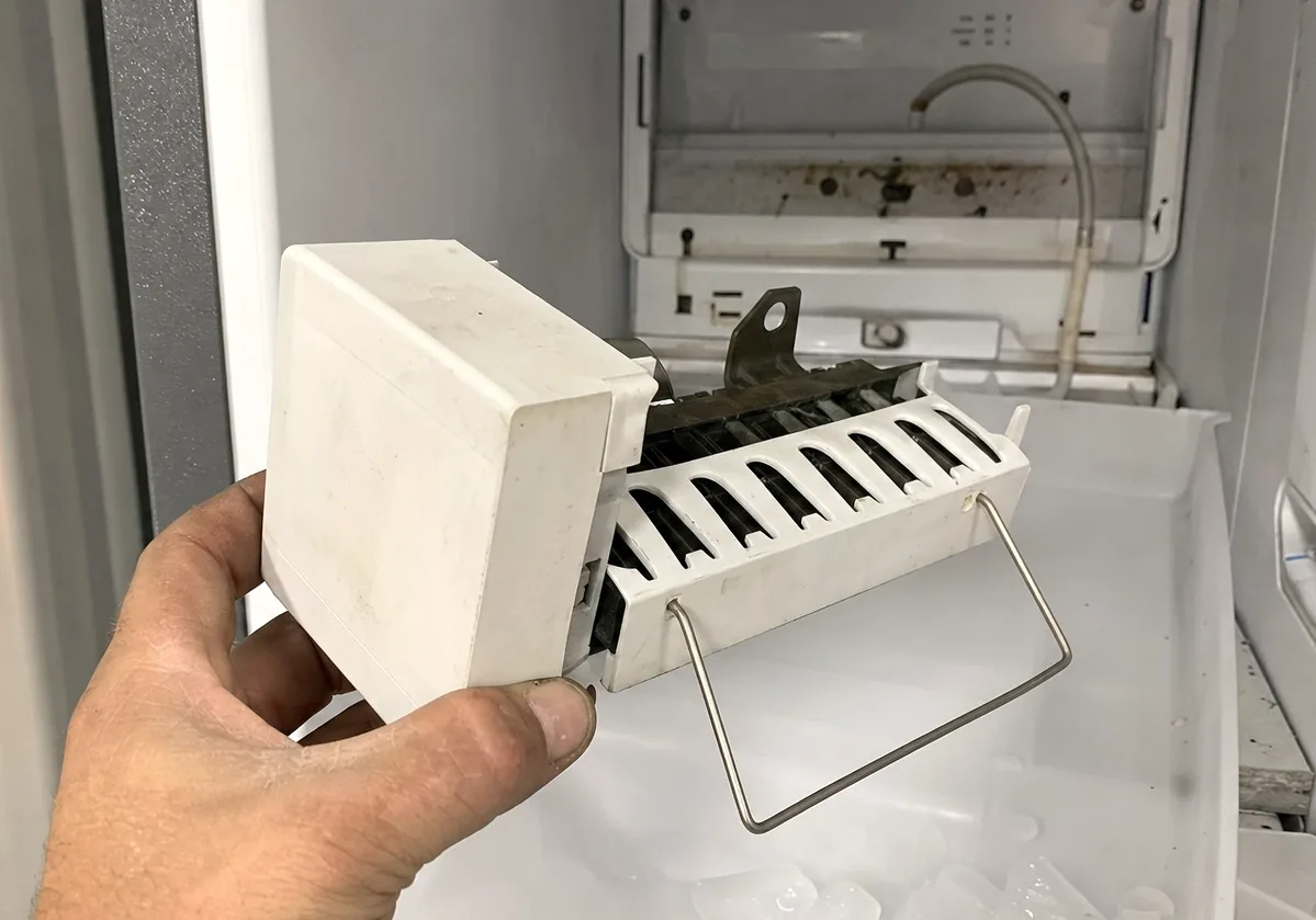 A technician holding an ice maker module beside the freezer bin it feeds, half-melted cubes heaped underneath.