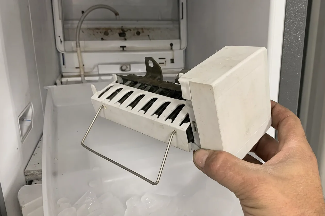 An ice maker module in a hand, the freezer door open behind it on shelves and a part-filled ice bin.