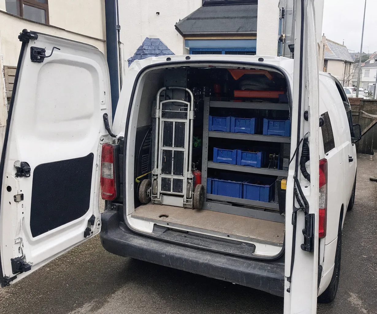 A white van on the wide new driveway of a recently built home, rear doors open on a folded hand truck and stock bins.